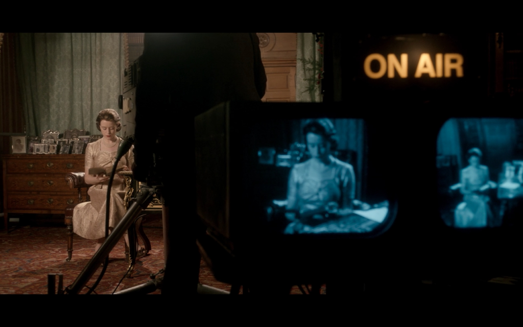Imagem da série The Crown, câmera filmando a Rainha com o sinal de ao vivo na tela e, ao fundo, imagem da rainha com livro na mão.